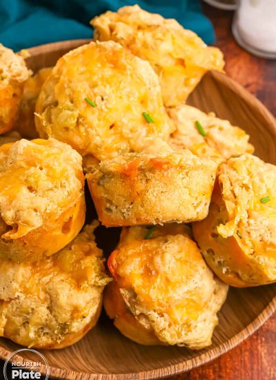 Close up of beer cheese bread muffins piled on a wooden plate, topped with melted cheddar and bits of green chile and chives.