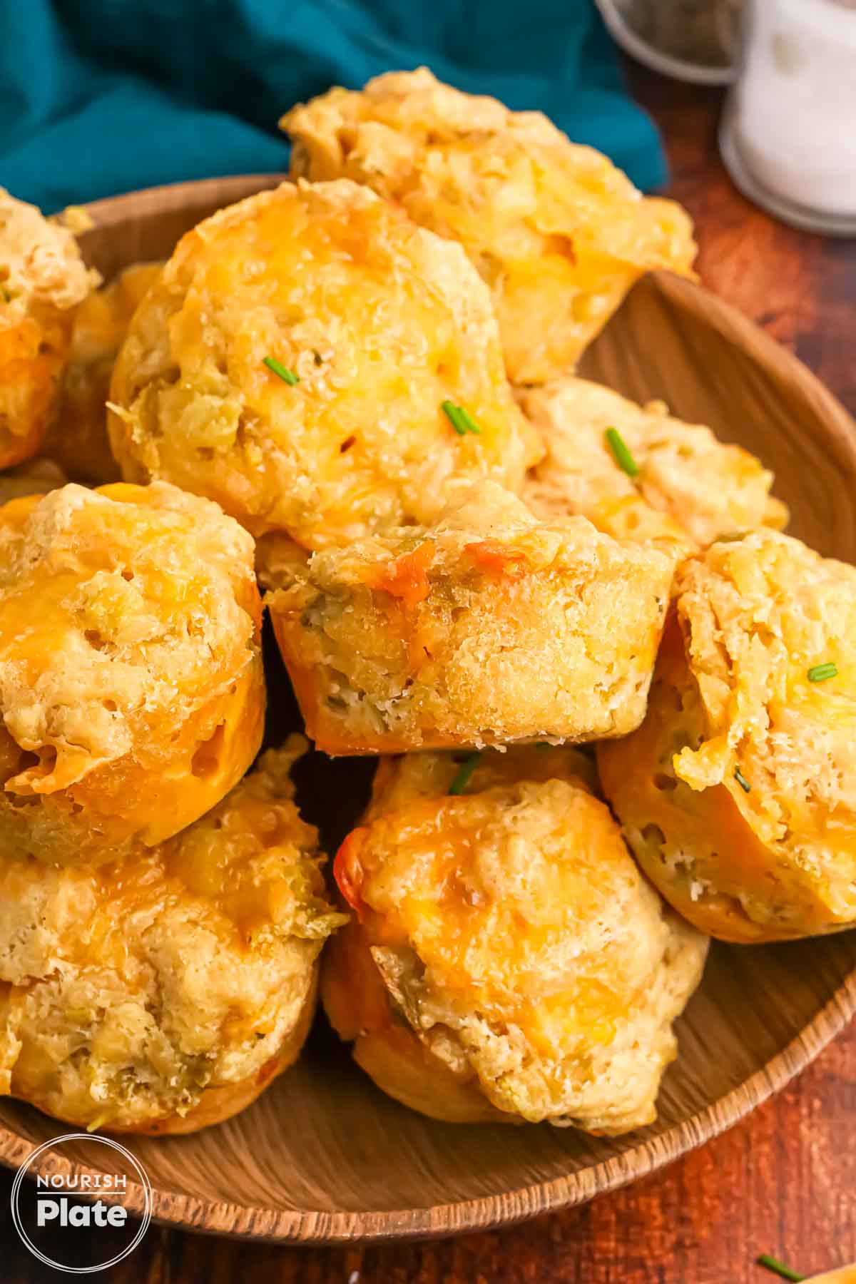 Close up of beer cheese bread muffins piled on a wooden plate, topped with melted cheddar and bits of green chile and chives.
