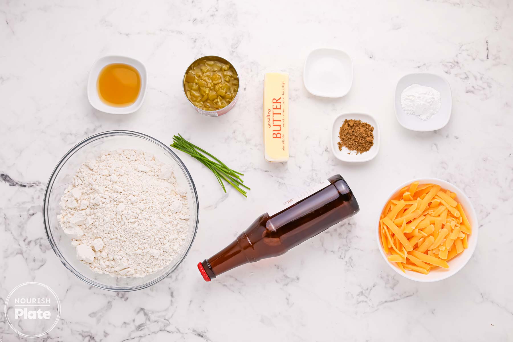 Ingredients for beer cheese bread muffins arranged on a white surface, including flour, beer, unsalted butter, honey, diced green chiles, cheddar cheese, chives, baking powder, salt, and ground cumin.