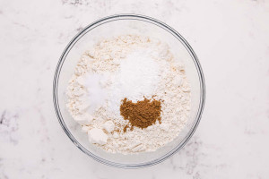Flour, baking powder, salt, and ground cumin added to a glass mixing bowl on a white surface.