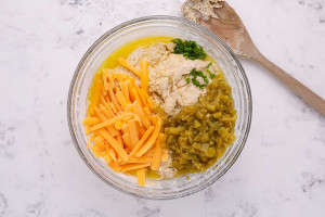 Muffin batter in a glass bowl topped with melted butter, shredded cheddar, diced green chiles, and chopped chives.