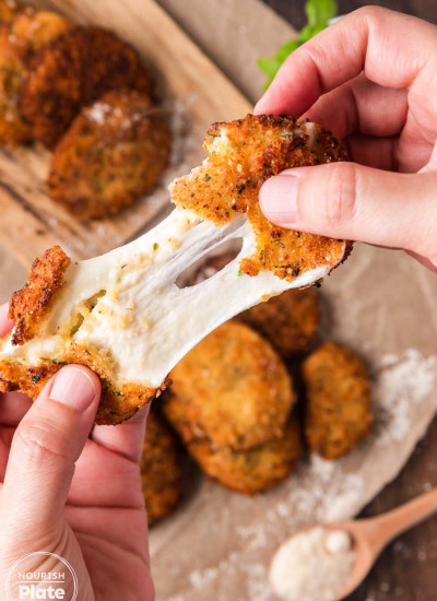 Close up of a fried mozzarella cheese slice being pulled apart to show the melted stretchy cheese inside, with more crispy breaded mozzarella slices blurred in the background.