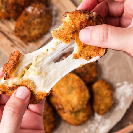 Close up of a fried mozzarella cheese slice being pulled apart to show the melted stretchy cheese inside, with more crispy breaded mozzarella slices blurred in the background.