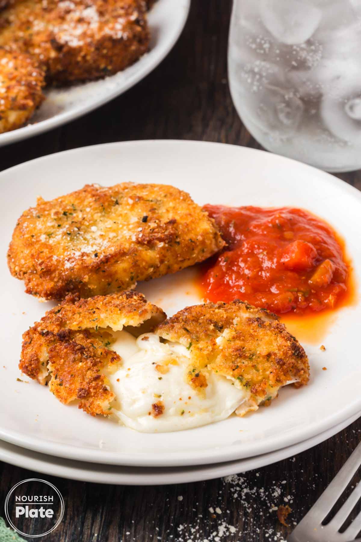 Fried mozzarella cheese slices on a plate with marinara sauce, including one cut open to show the melted cheese center, with more breaded mozzarella in the background.