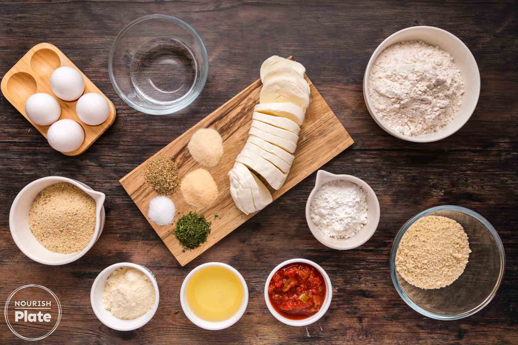 Overhead view of ingredients for fried mozzarella cheese slices, including sliced fresh mozzarella, eggs, flour, cornstarch, panko, Italian style breadcrumbs, Parmesan cheese, dried herbs, seasonings, oil, and marinara sauce on a dark wooden surface.