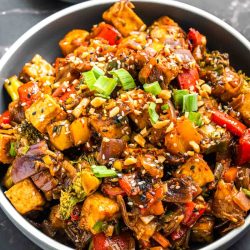 A bowl of crispy tofu stir fry with broccoli, red and yellow bell peppers, red onion, and carrot in a glossy dark sauce, topped with chopped peanuts, sesame seeds, and sliced green onions.
