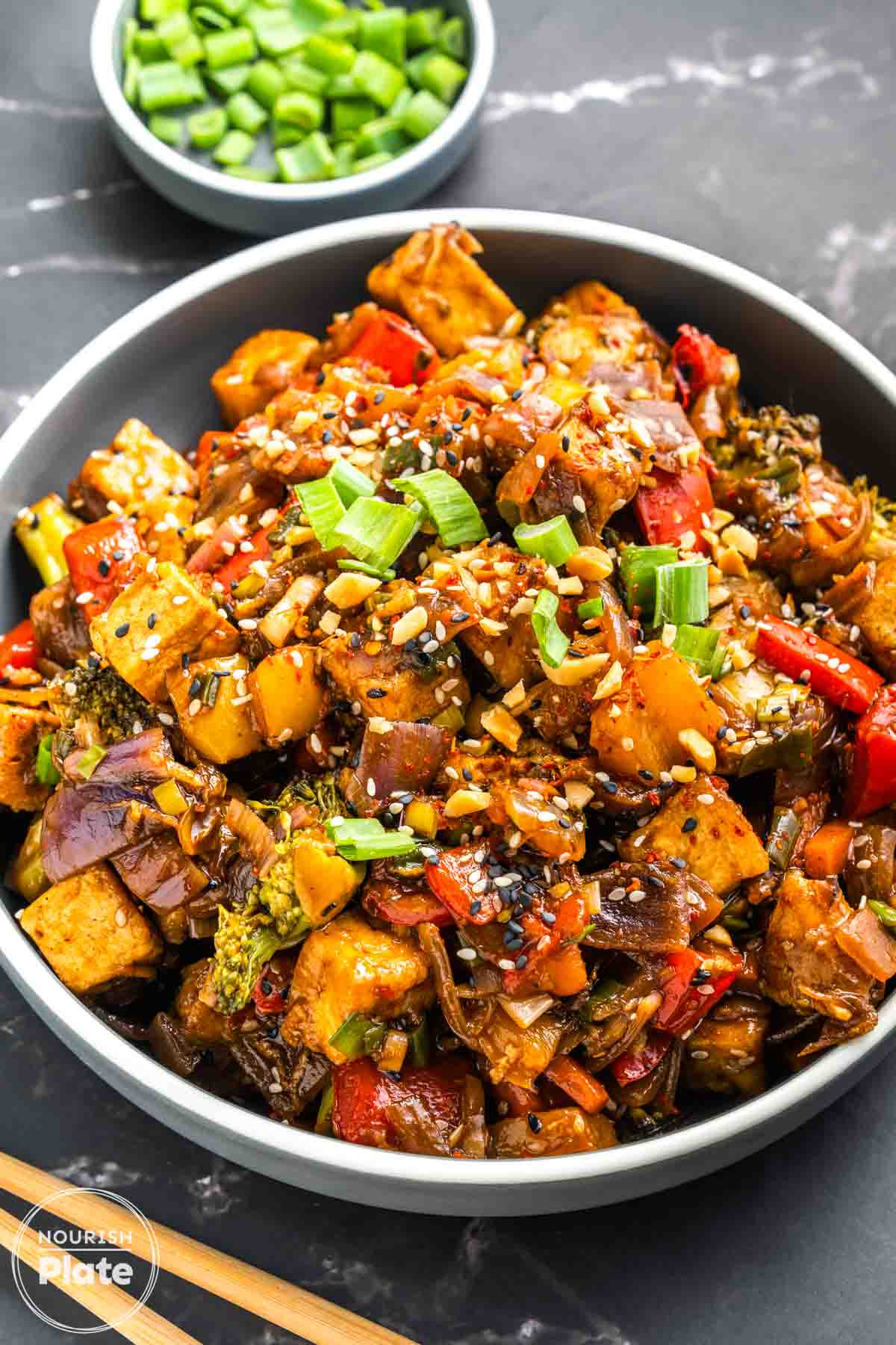 A bowl of crispy tofu stir fry with broccoli, red and yellow bell peppers, red onion, and carrot in a glossy dark sauce, topped with chopped peanuts, sesame seeds, and sliced green onions.