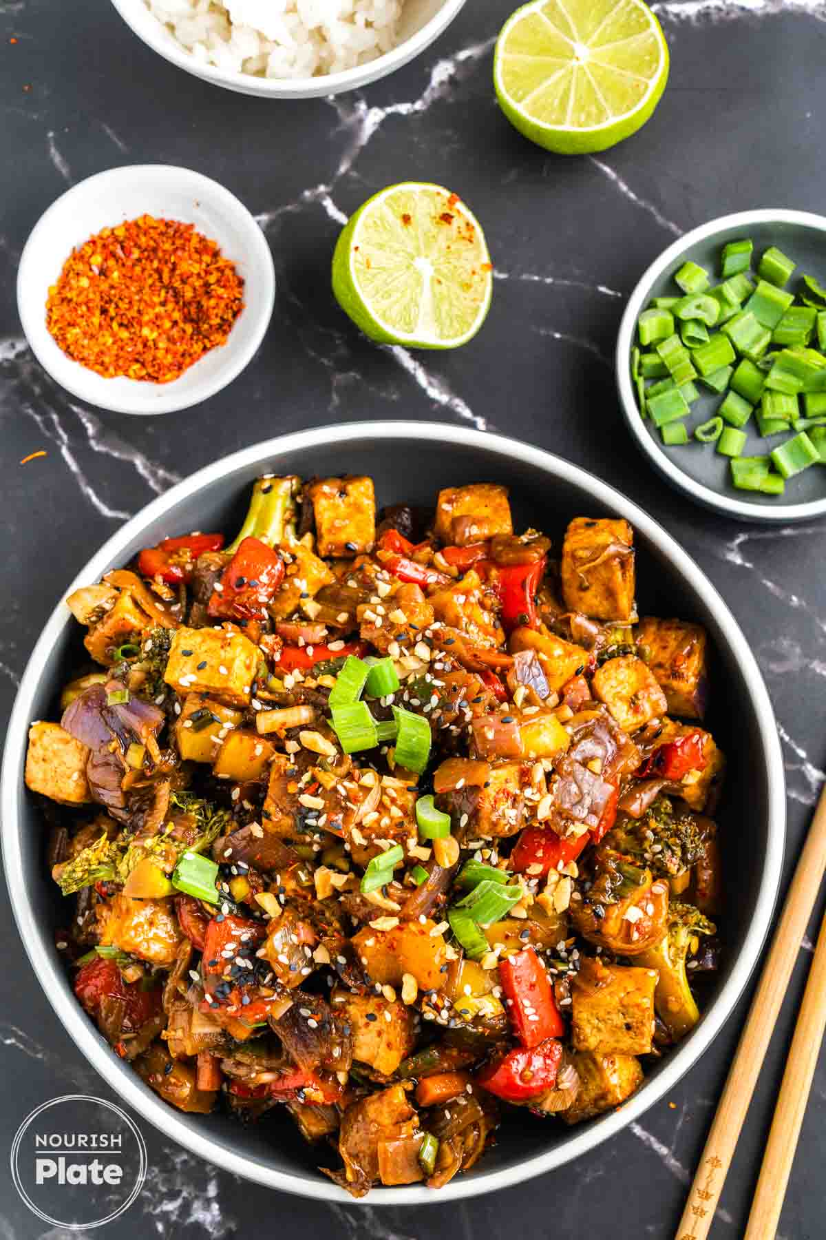 Overhead view of crispy tofu stir fry in a bowl with broccoli, red and yellow bell peppers, and red onion in a glossy sauce, topped with green onions, sesame seeds, and chopped peanuts, with lime, chili flakes, and chopsticks on the side.