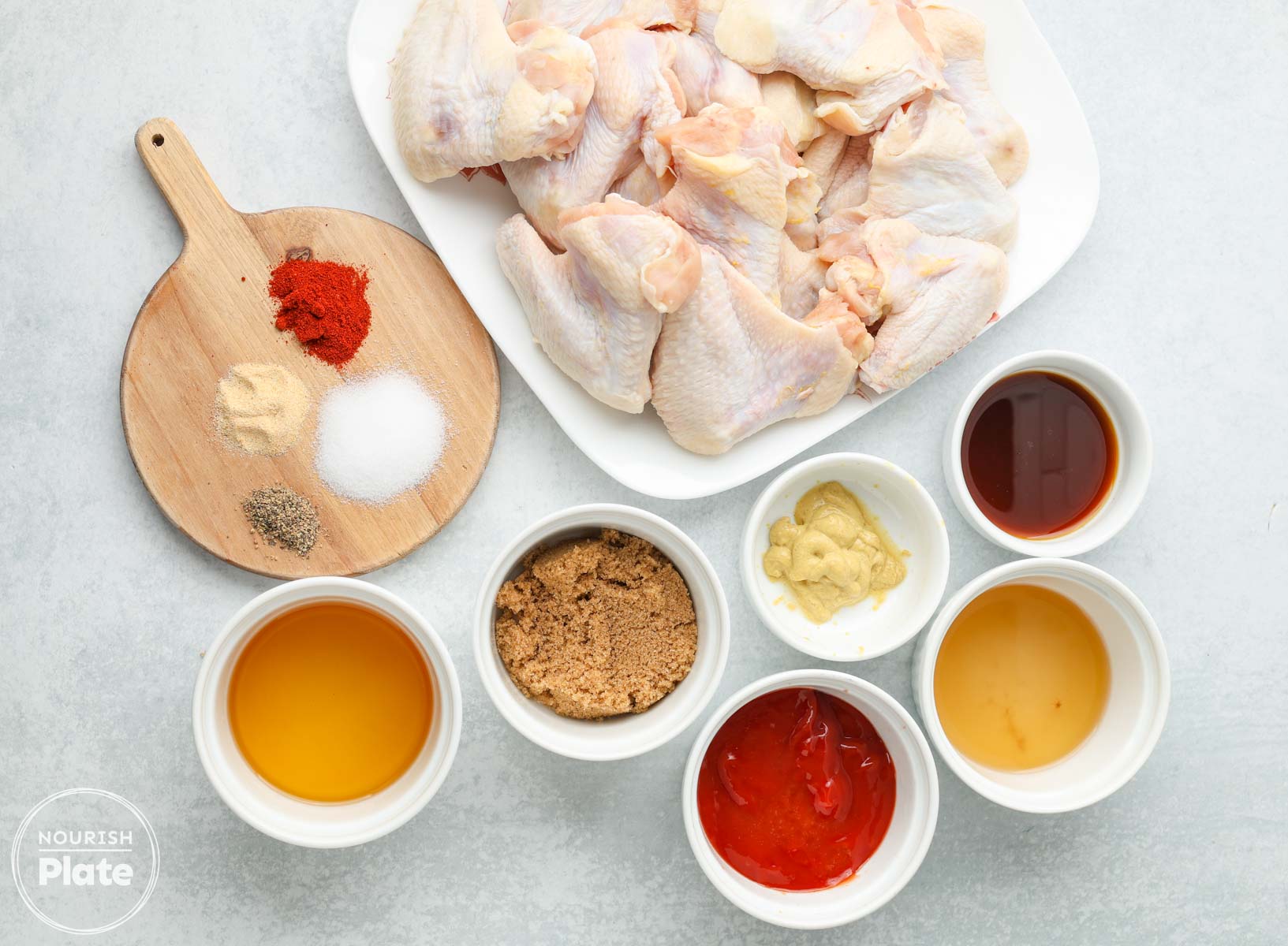 Ingredients for baked BBQ chicken wings arranged on a countertop, including raw chicken wings, BBQ sauce, honey, mustard, brown sugar, vinegar, oil, and seasonings.