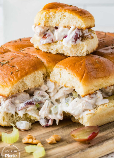 Close up of chicken salad sliders on Hawaiian rolls with grapes, celery, walnuts, and thyme on a wooden board.