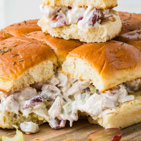 Close up of chicken salad sliders on Hawaiian rolls with grapes, celery, walnuts, and thyme on a wooden board.