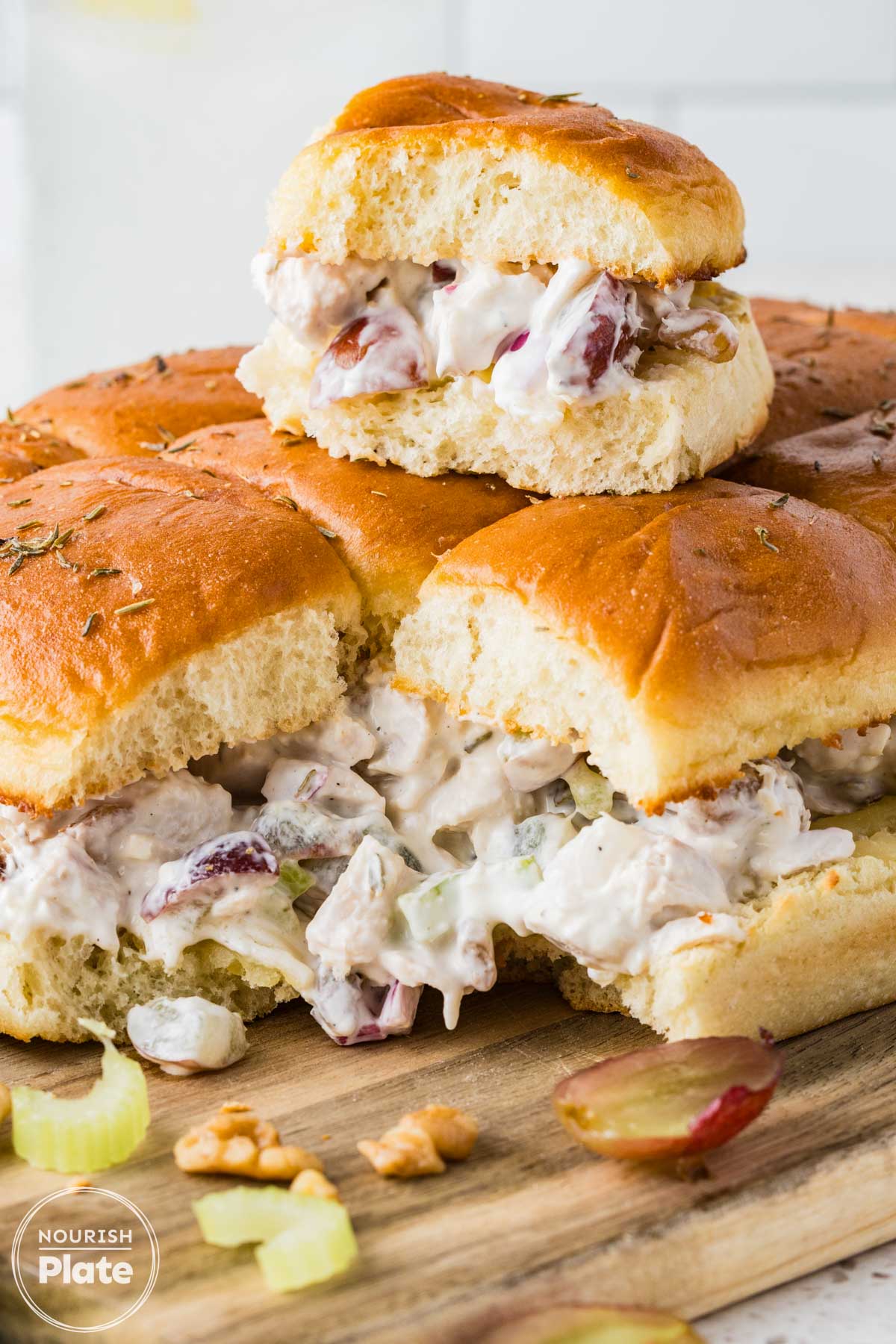 Close up of chicken salad sliders on Hawaiian rolls with grapes, celery, walnuts, and thyme on a wooden board.