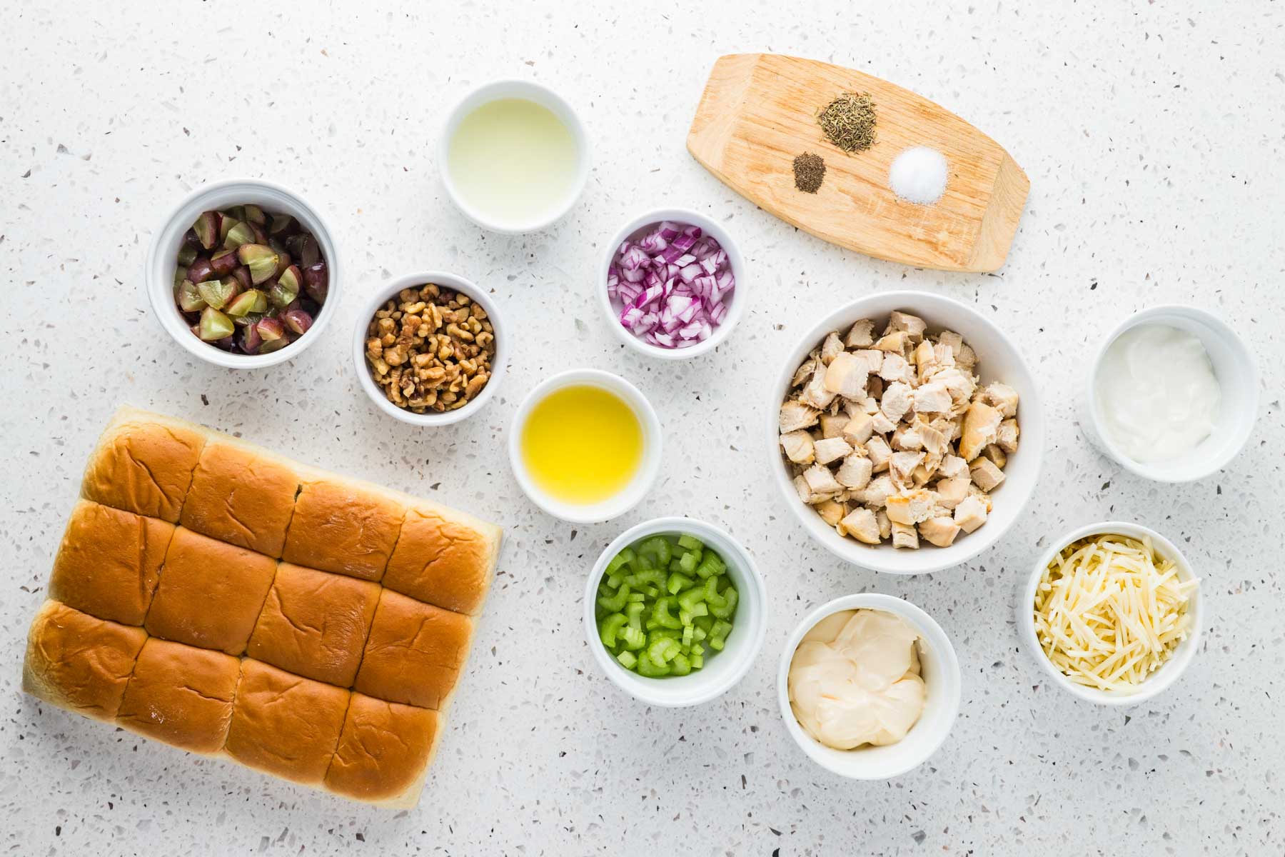 Ingredients for chicken salad sliders arranged on a countertop, including slider rolls, chicken, grapes, walnuts, celery, onion, cheese, mayonnaise, sour cream, oil, lemon juice, and seasonings.