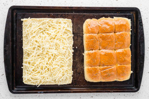 Split slider rolls on a baking sheet with shredded cheese on the bottom half and seasoned tops beside it.