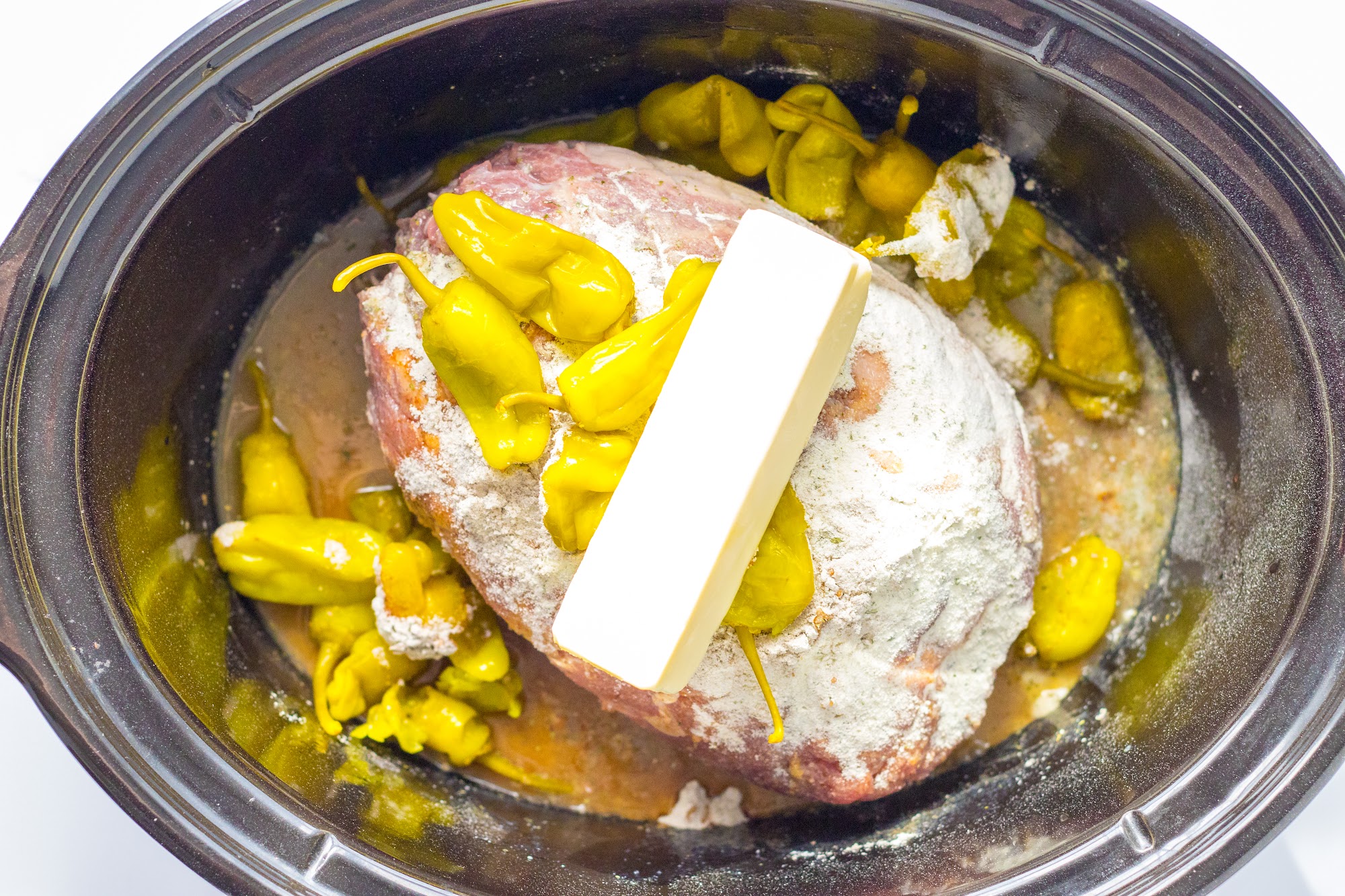 Pork roast in a slow cooker topped with ranch and gravy mix, a stick of butter, and pepperoncini