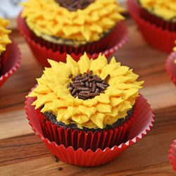 Sunflower cupcakes with yellow buttercream petals and chocolate sprinkle centers in red liners on a wooden board