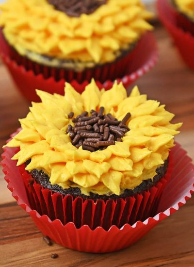 Sunflower cupcakes with yellow buttercream petals and chocolate sprinkle centers in red liners on a wooden board