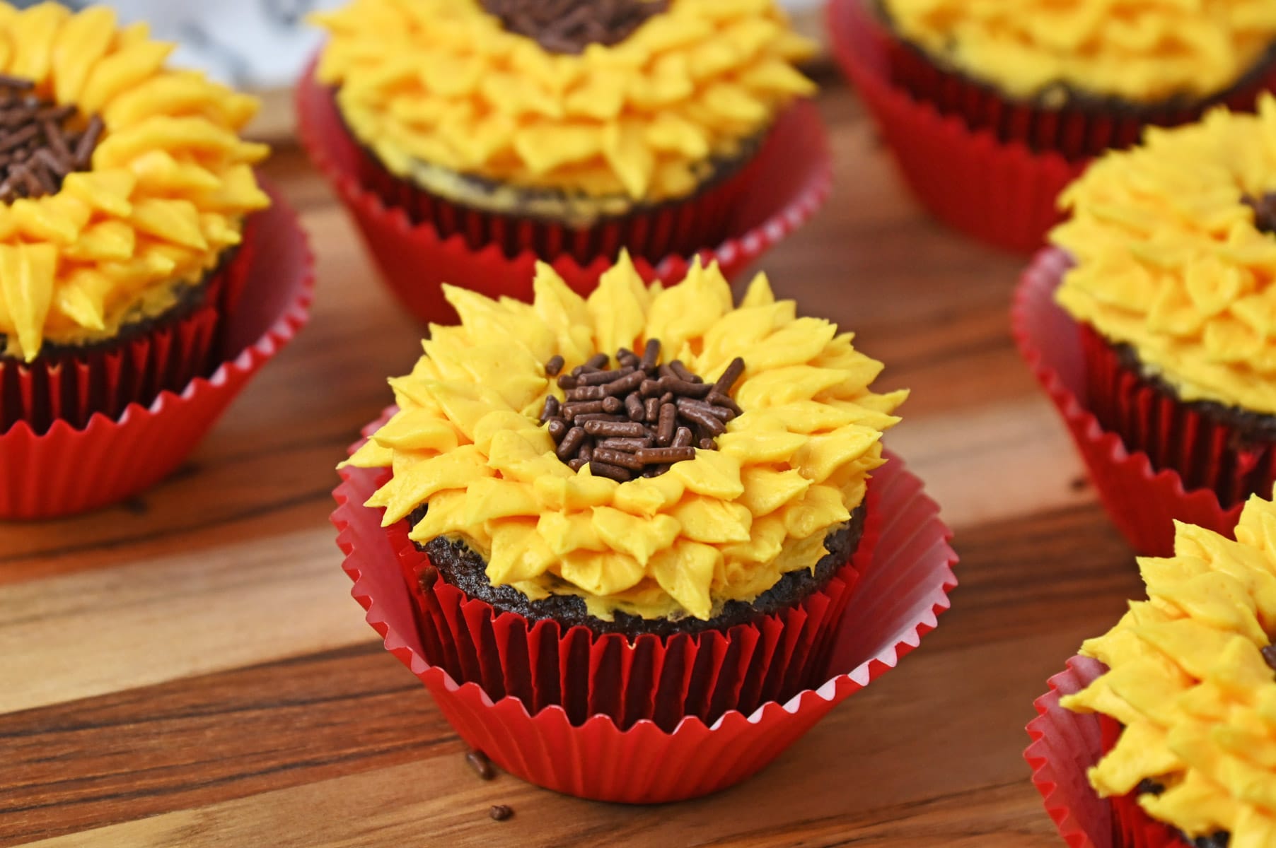 Sunflower cupcakes with yellow buttercream petals and chocolate sprinkle centers in red liners on a wooden board