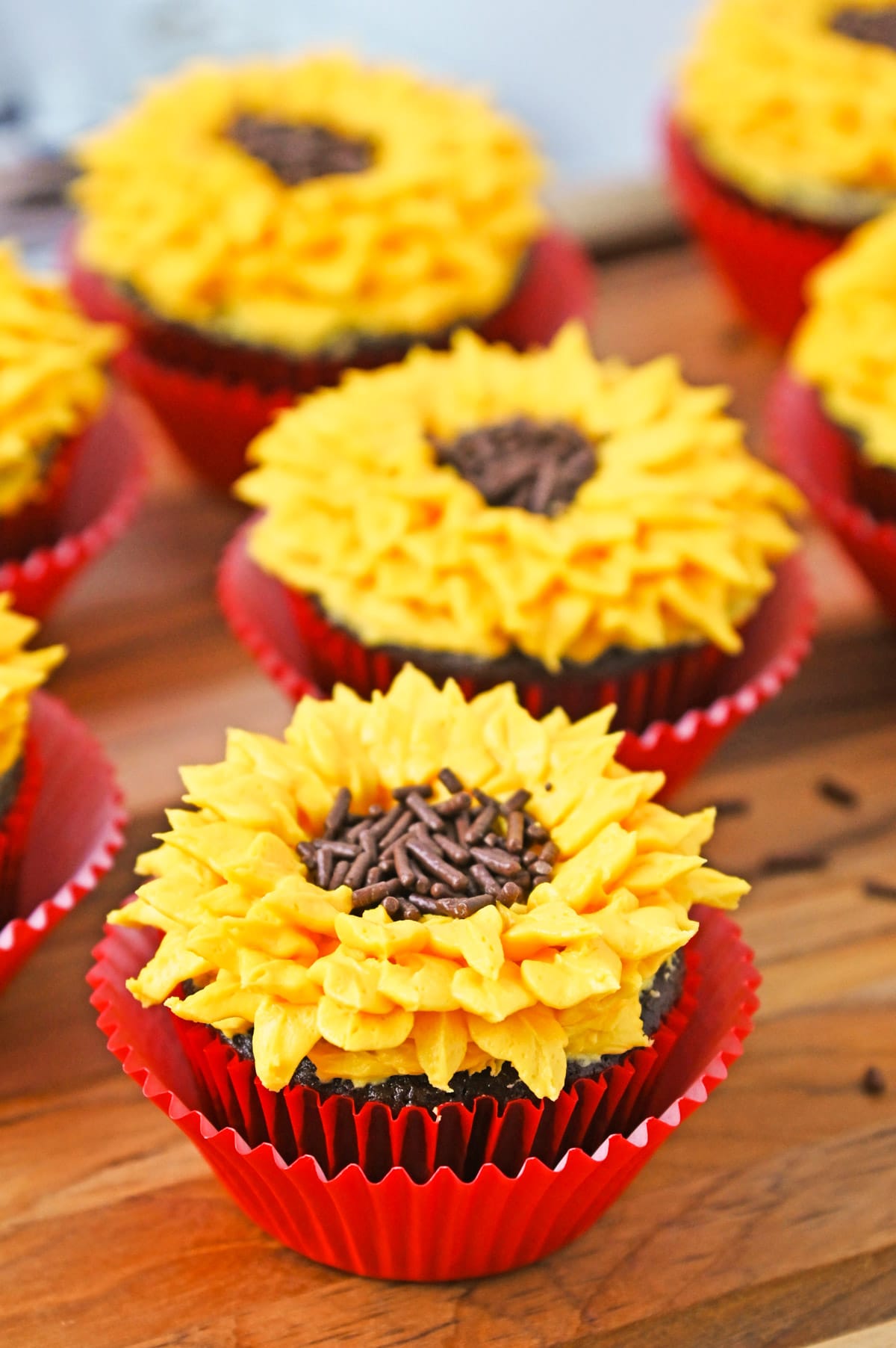 Sunflower cupcakes in red liners on a wooden board topped with yellow frosting petals and chocolate sprinkles