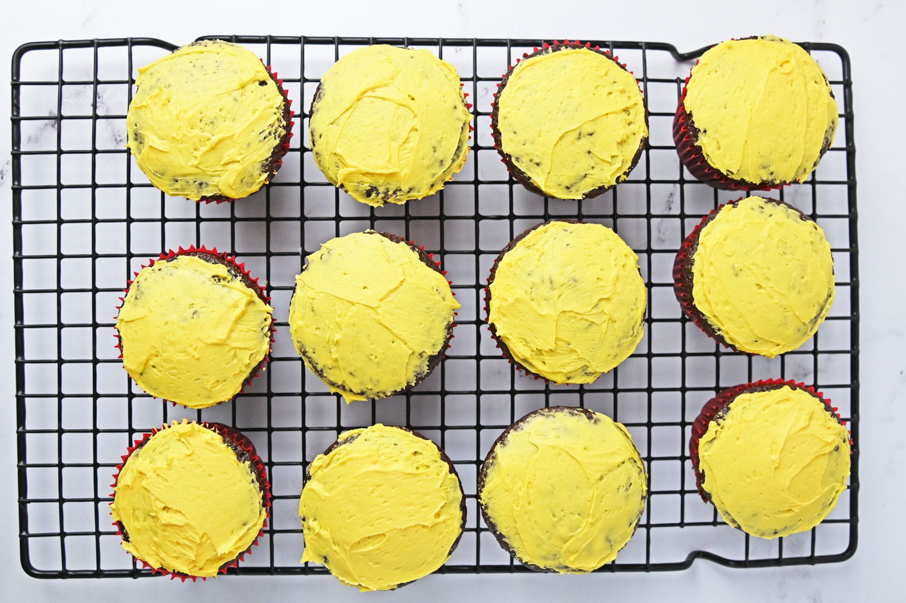 Twelve chocolate cupcakes on a wire rack topped with a thin layer of yellow frosting